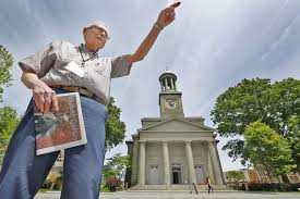 starszy pan w okularach wskauje palce w górę. W tle widać zabytkowy budynek