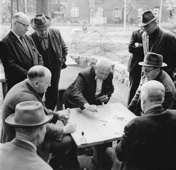 mężczyźni zgronadzeni na dworze wokół stolika. niektórzy siedzą i grają w karty - w skat, inni sie przygladaja. 