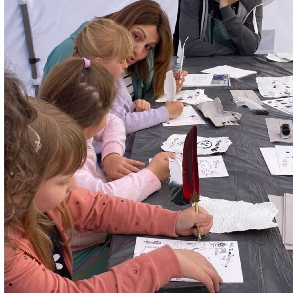 grupa osób piszących gęsim piórem. Przed sobą mają karki z napisami