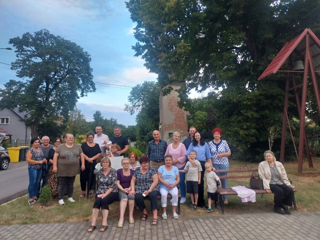 mieszkancywsi, kobiety, mężczyźni i dzieci. Zgdr5omadzeni na placu, przy wieży z dzwonem. niektórzy siedzą, niektórzy stoją.
