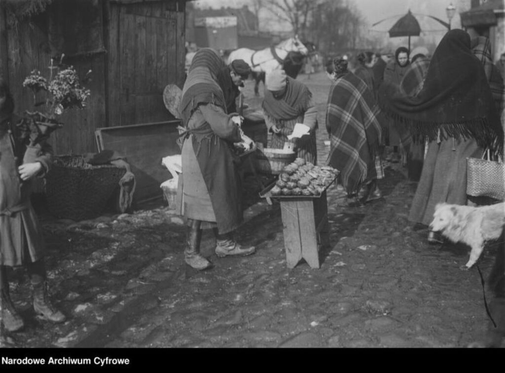 klepisko, wieś albo jakieś małe stare miasrto, w tle drewniane domy, na ulicy ludzie handlują. na pierwszym planie kobieta w chuście na płaszczu, spódnicy. Przed nią stoi stołek drewniany a na nim bl;acha z pączkami. W tle inni ludzie i koń. Zdjęcie czarnobiałe archiwalne